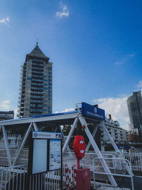 A view of Chelsea Harbour Pier, featuring a white metal structure with a blue sign displaying the pier's name and an information board mounted on a black frame. A red lifebuoy is attached to the struc