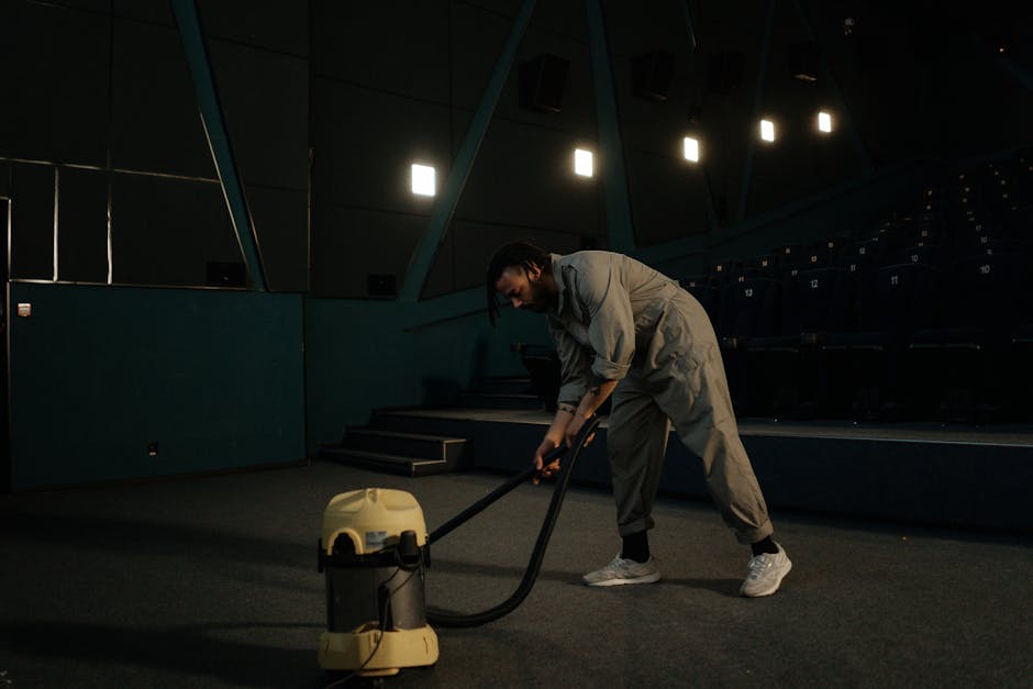 A person performing deep cleaning on a carpeted floor in a darkened room, likely a commercial or conference space. The individual is using a long-handled cleaning tool, possibly for sanitisation or du