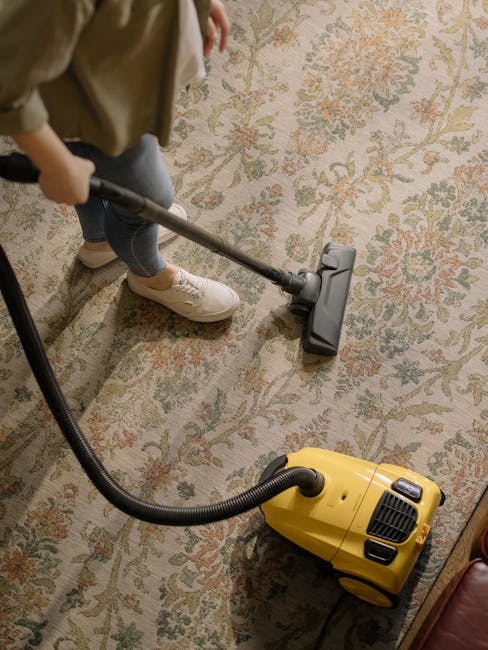 A yellow canister vacuum cleaner with a flexible hose and cleaning head is positioned on an ornate, patterned area rug with floral design in shades of beige, green, and orange, placed on a wooden floo
